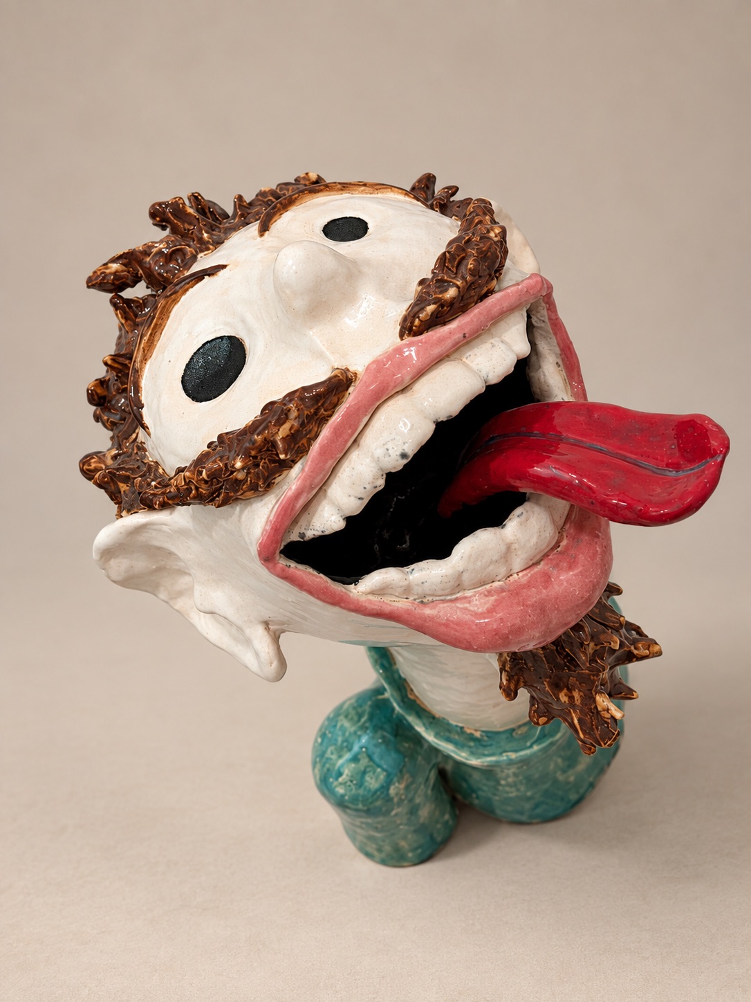Ceramic self-portrait by Spencer Tracy Brown with an exaggerated open mouth, extended tongue, simplified black eyes, textured brown hair, and turquoise body